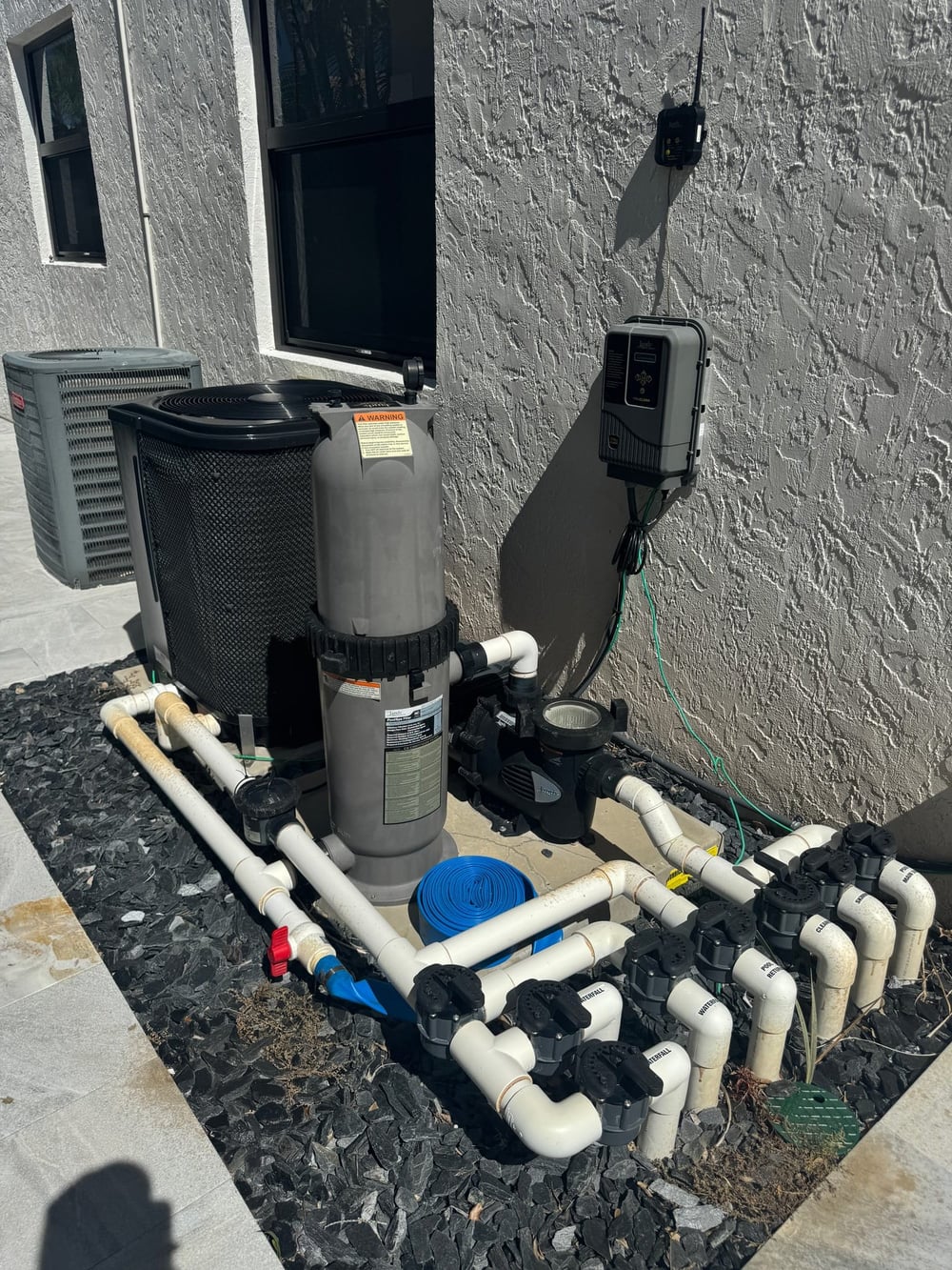 A South Florida pool equipment pad with a heat pump behind a cartridge filter, a variable-speed pump, valve manifold, and a wall-mounted automation panel with a wireless antenna